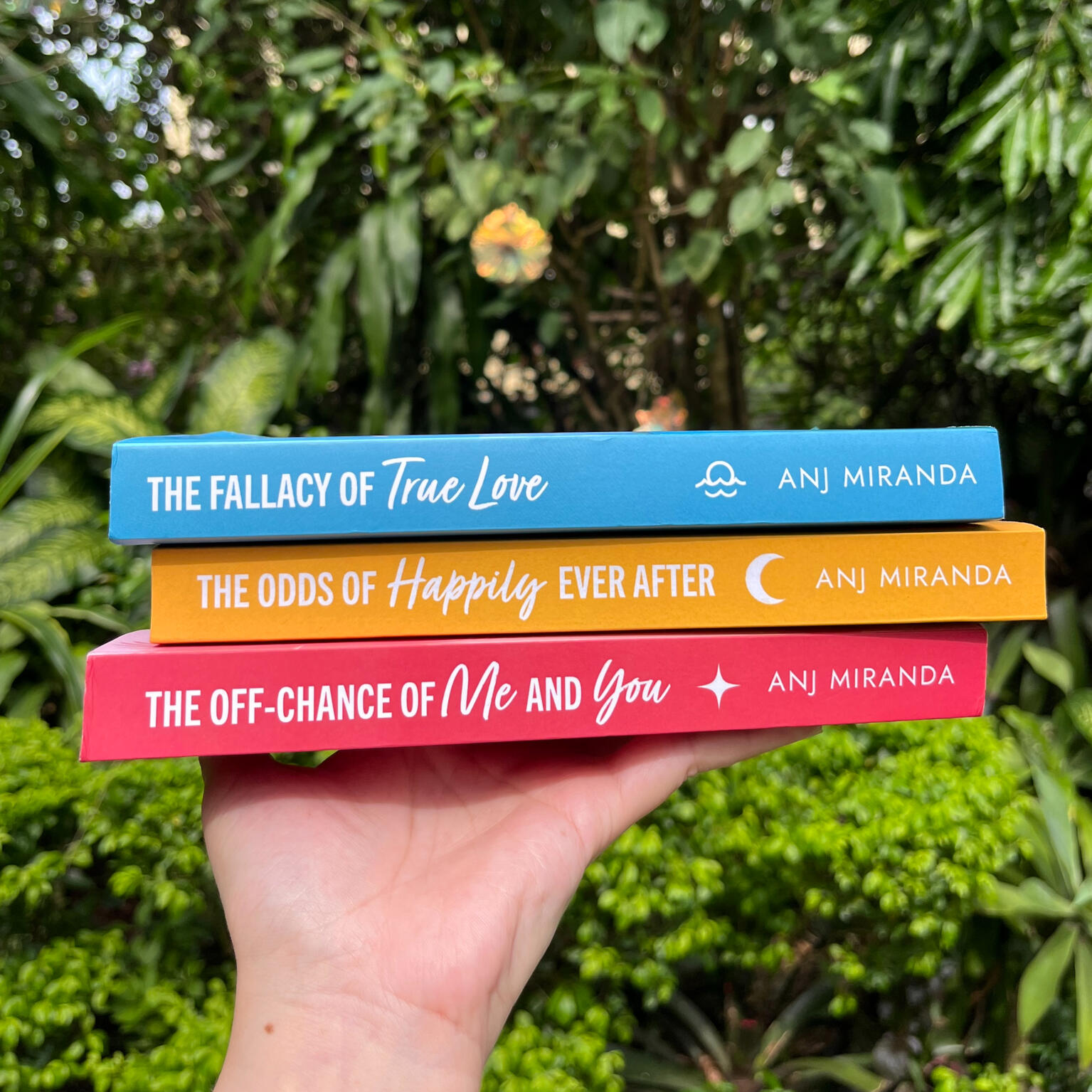 hand holding paperback books of the fallacy of true love, the odds of happily ever after, and the off-chance of me and you by anj miranda